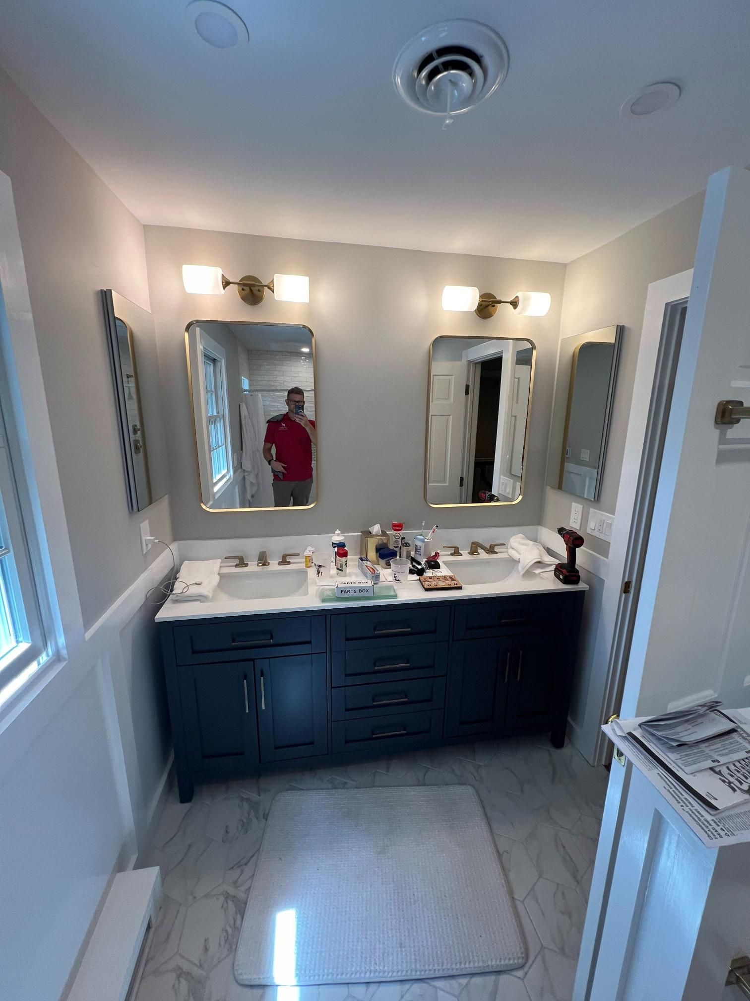 Bright bathroom with double vanity, mirrors, sconces, and a person in a red shirt reflected in the mirror.