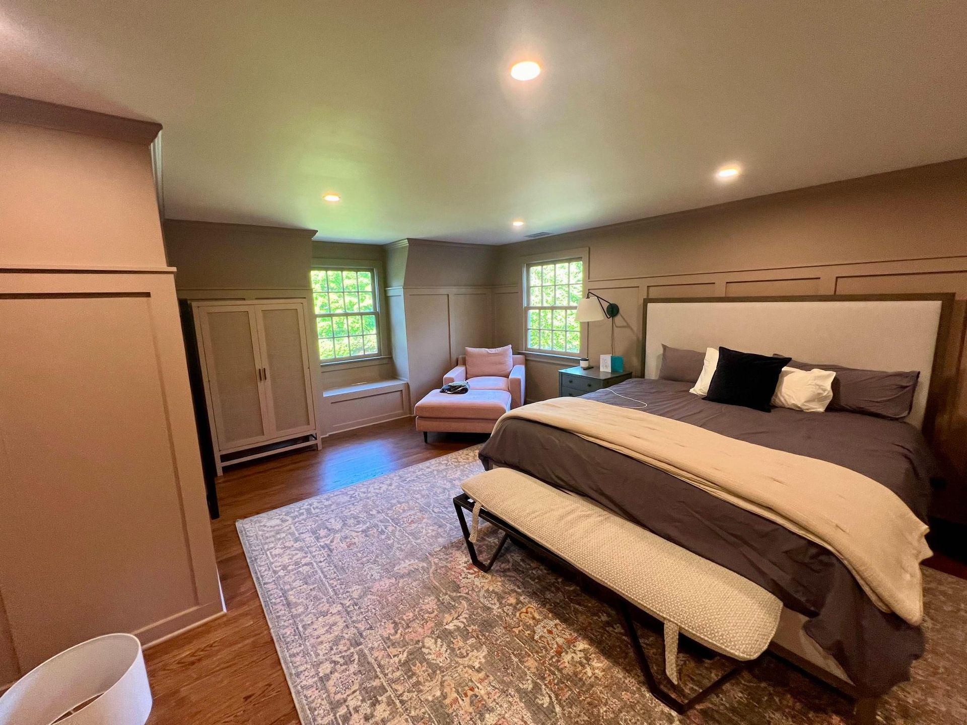 Spacious bedroom with a large bed, bench, armoire, armchair, and two windows.