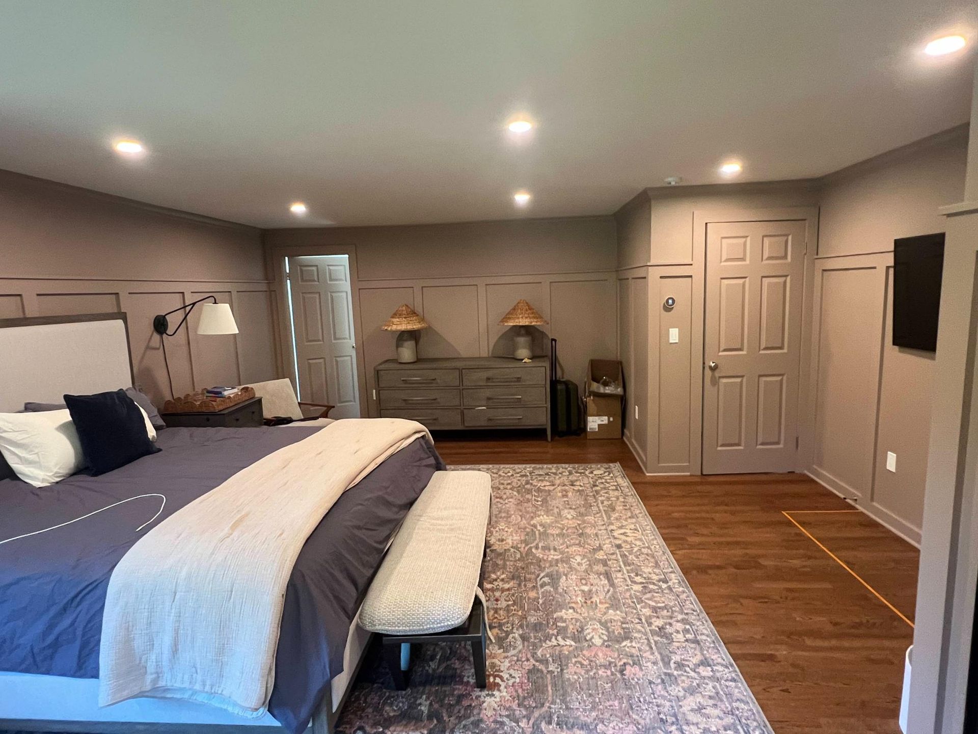 Spacious bedroom with a king bed, bench, dresser, gray walls, hardwood floor, and wall-mounted TV.