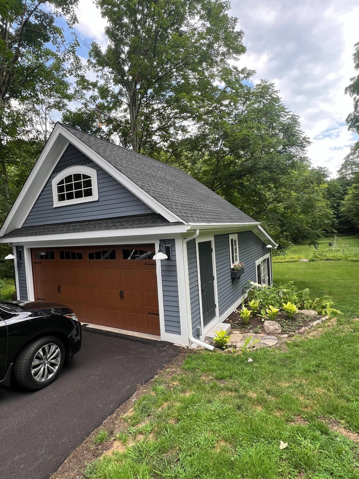 Gray house with detached garage, white trim, brown door, black car, and green lawn under trees
