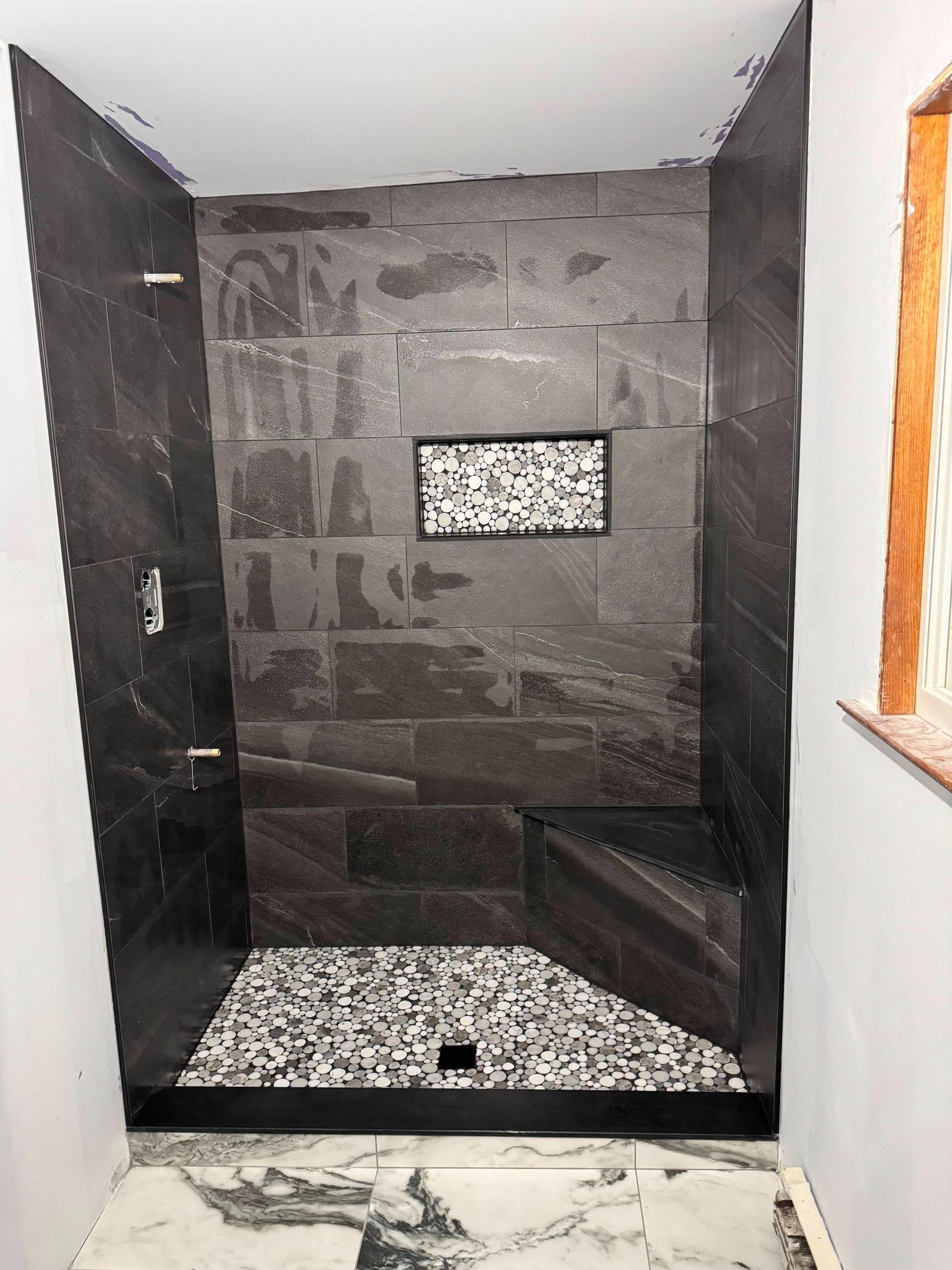 Modern tiled shower stall with black trim, pebble floor, and a small frosted window.