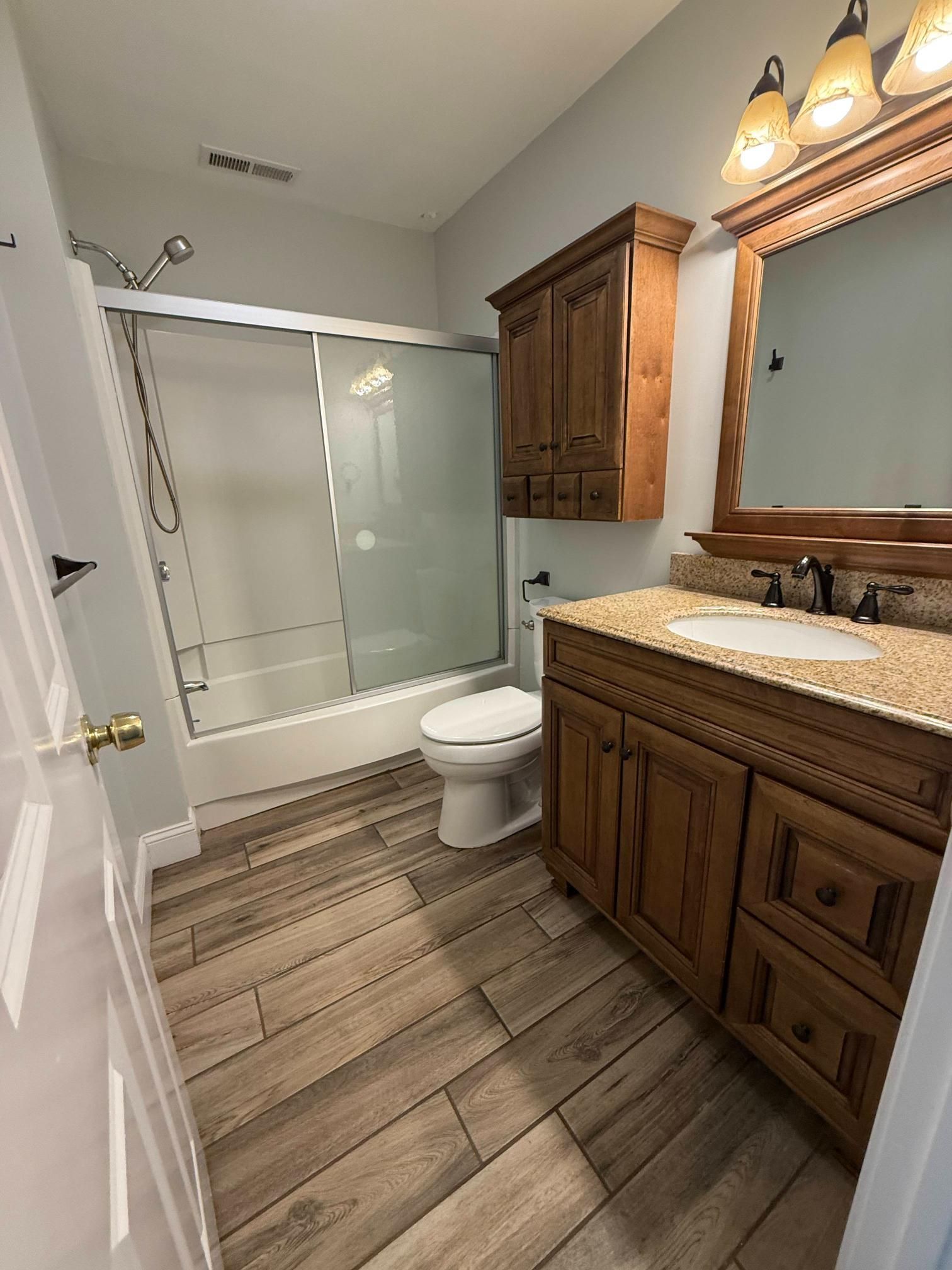 Bathroom with wood vanity, large mirror, tub-shower, toilet, and wood-look tile floor.