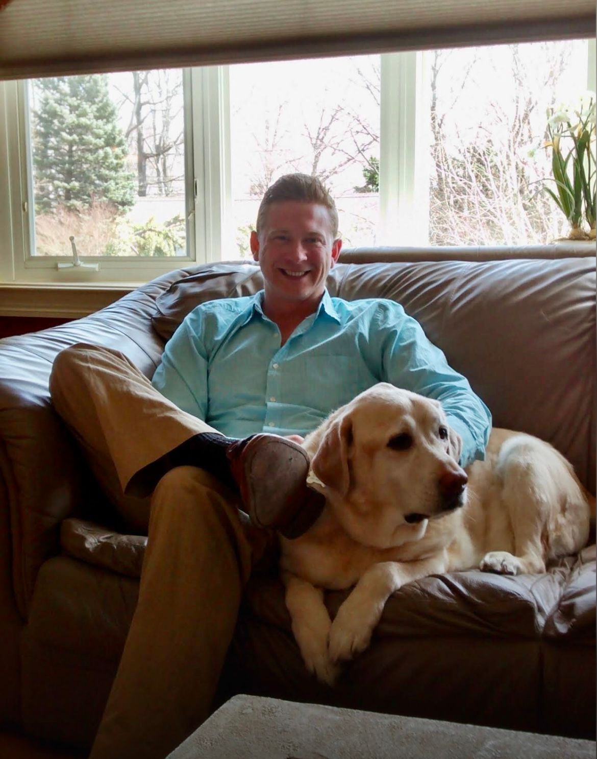 A man sits on a leather couch with a golden retriever, in front of large windows.