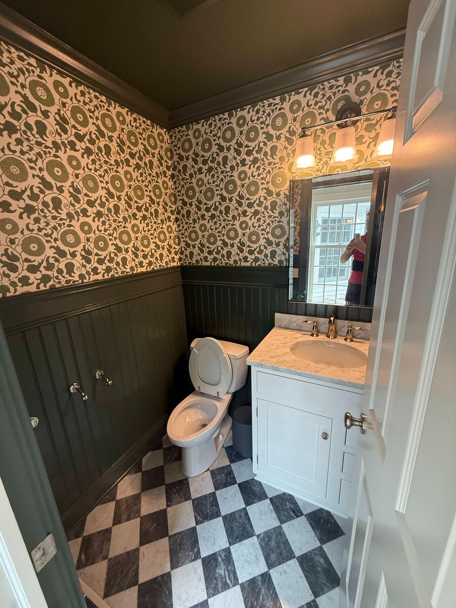 Small bathroom with black-and-white checkered floor, floral wallpaper, toilet, vanity, and mirror light.