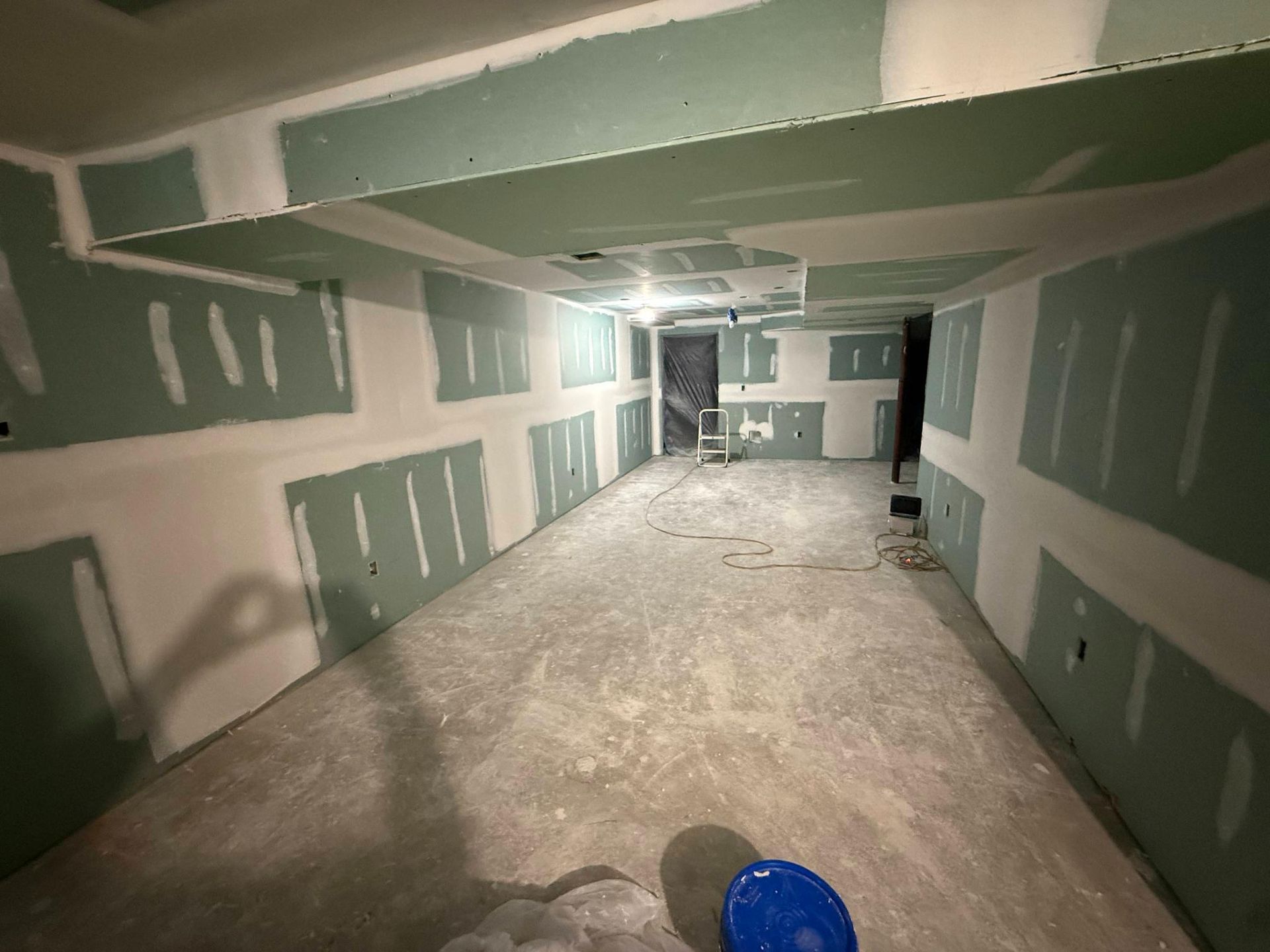 Unfinished drywall room under construction with taped seams, bare floor, and a blue bucket in the foreground