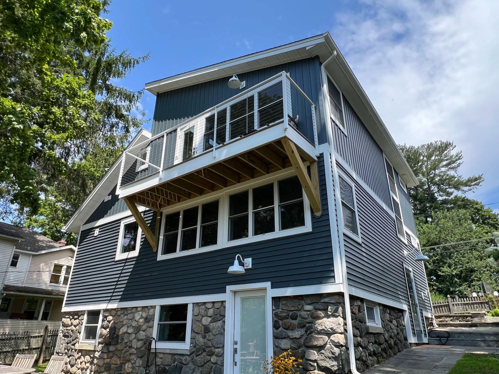 Dark blue two-story house with stone base, white trim, and a balcony under a bright sky