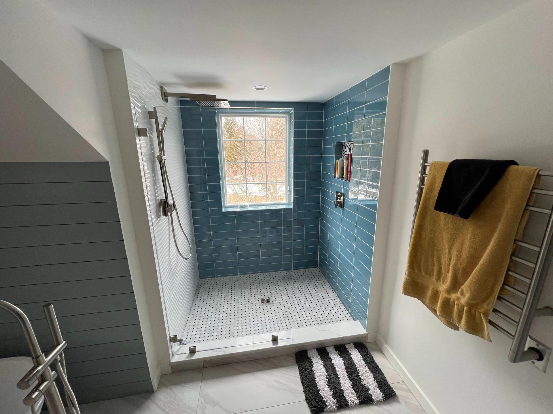 Small tiled walk-in shower with blue walls, window, showerhead, and towels hanging on the side.