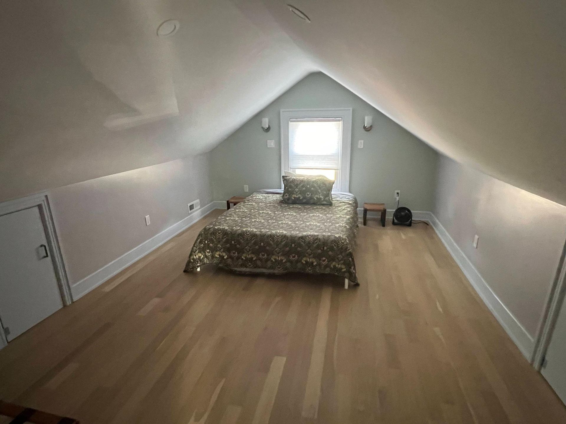 Attic bedroom with sloped white ceiling, wood floor, bed, and small window at the far end