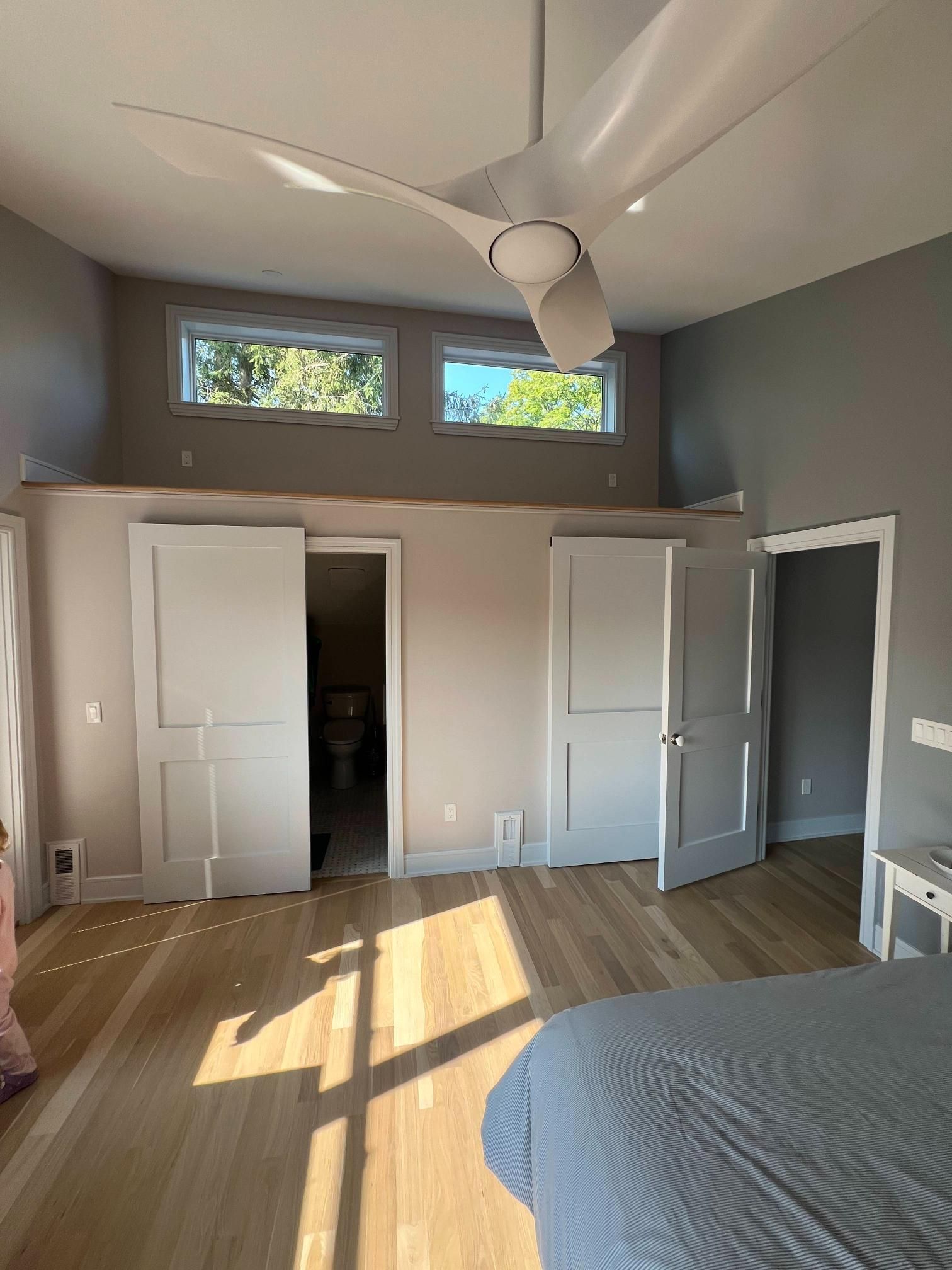 Sunlit bedroom with open closet doors, gray walls, wood floor, and a ceiling fan.