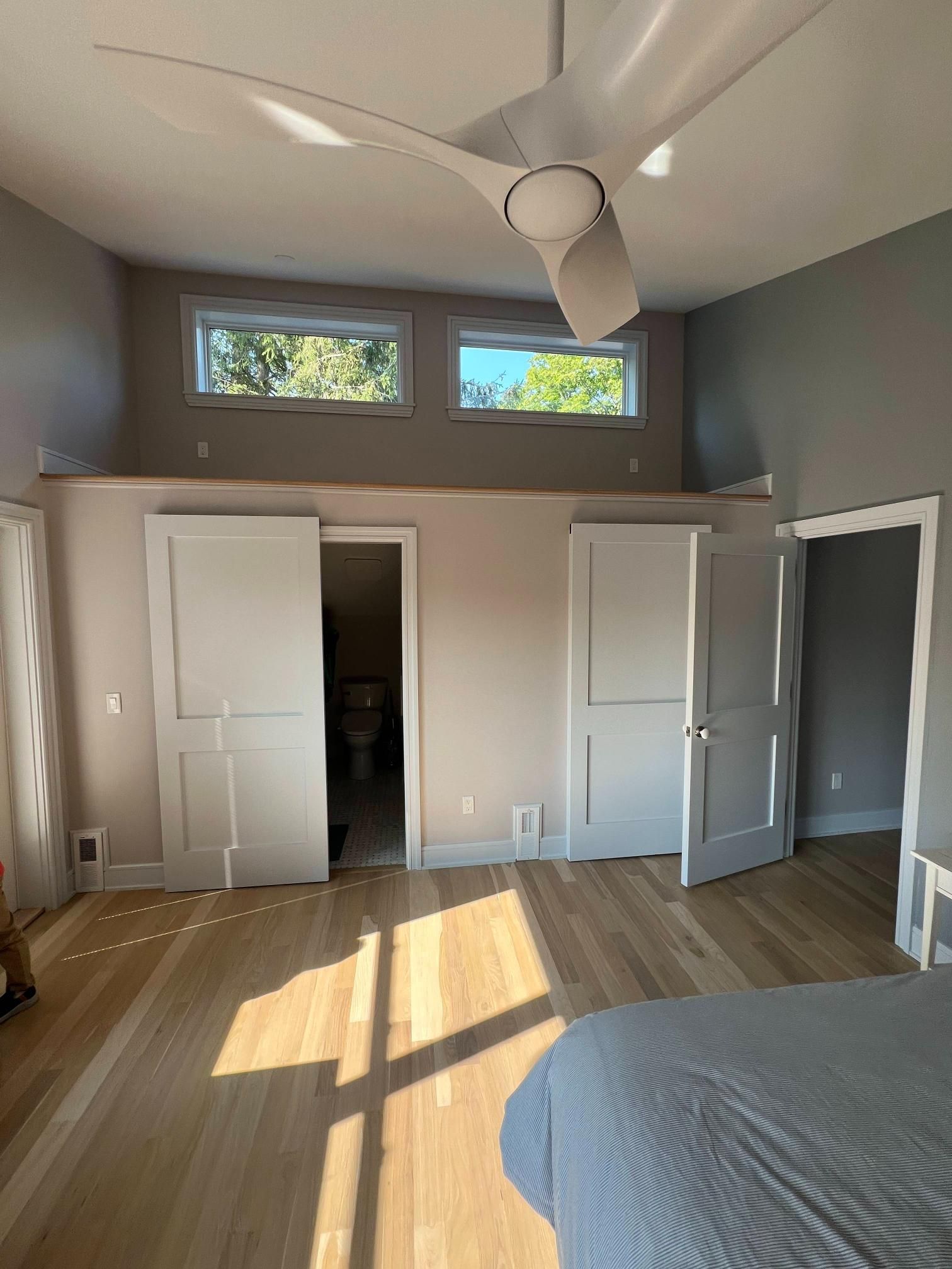 Bright bedroom with wood floors, gray walls, high windows, ceiling fan, open closet doors, and sunlight on the floor