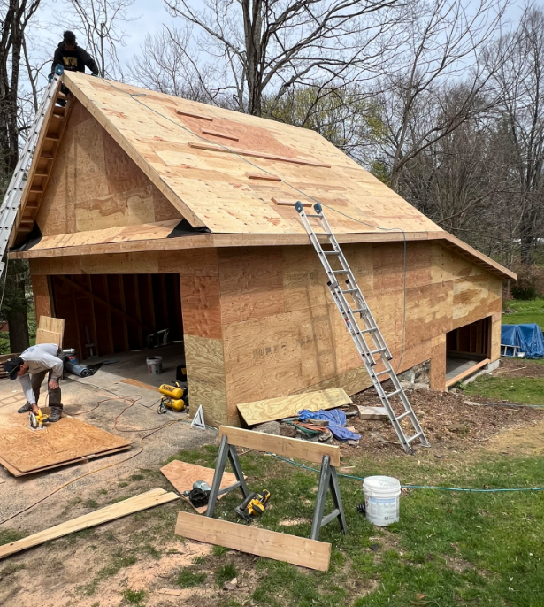 Workers installing a plywood roof on a brick garage, with ladders and tools on a grassy lot.
