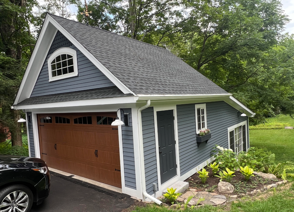 Blue garage with brown door, white trim, and dormer window beside a landscaped yard.