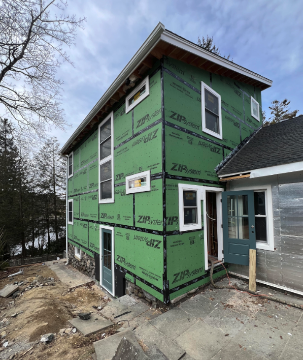 Green house under construction with wrapped exterior, windows installed, and porch beside a wooded yard