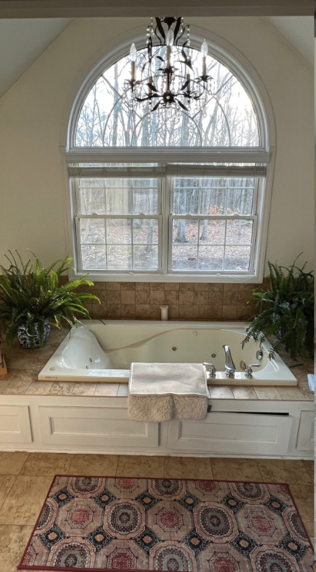 Bright bathroom with freestanding tub beneath arched window, chandelier, plants, and patterned rug.