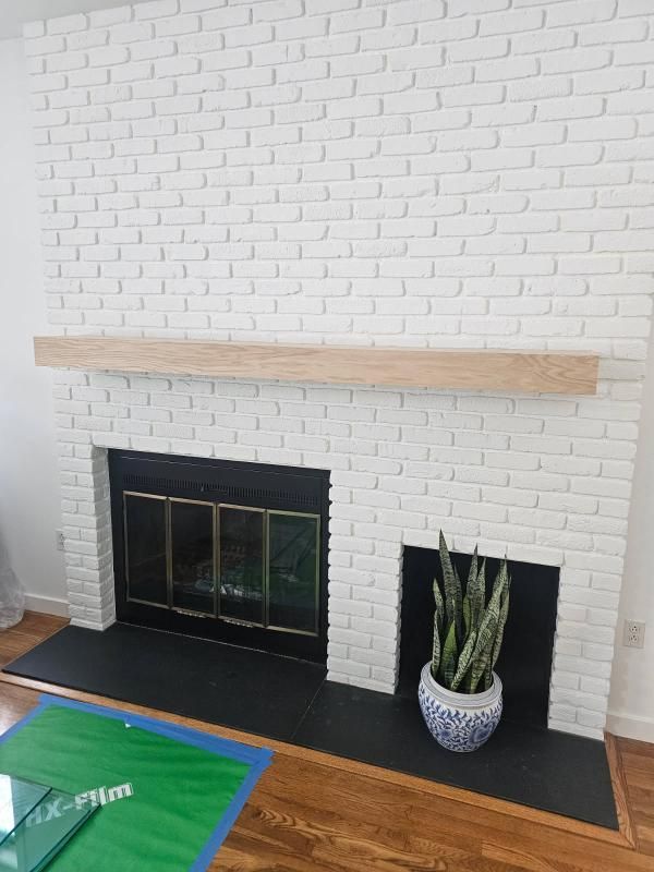 White brick fireplace with black firebox and hearth, wood mantel, and potted plant on the right.