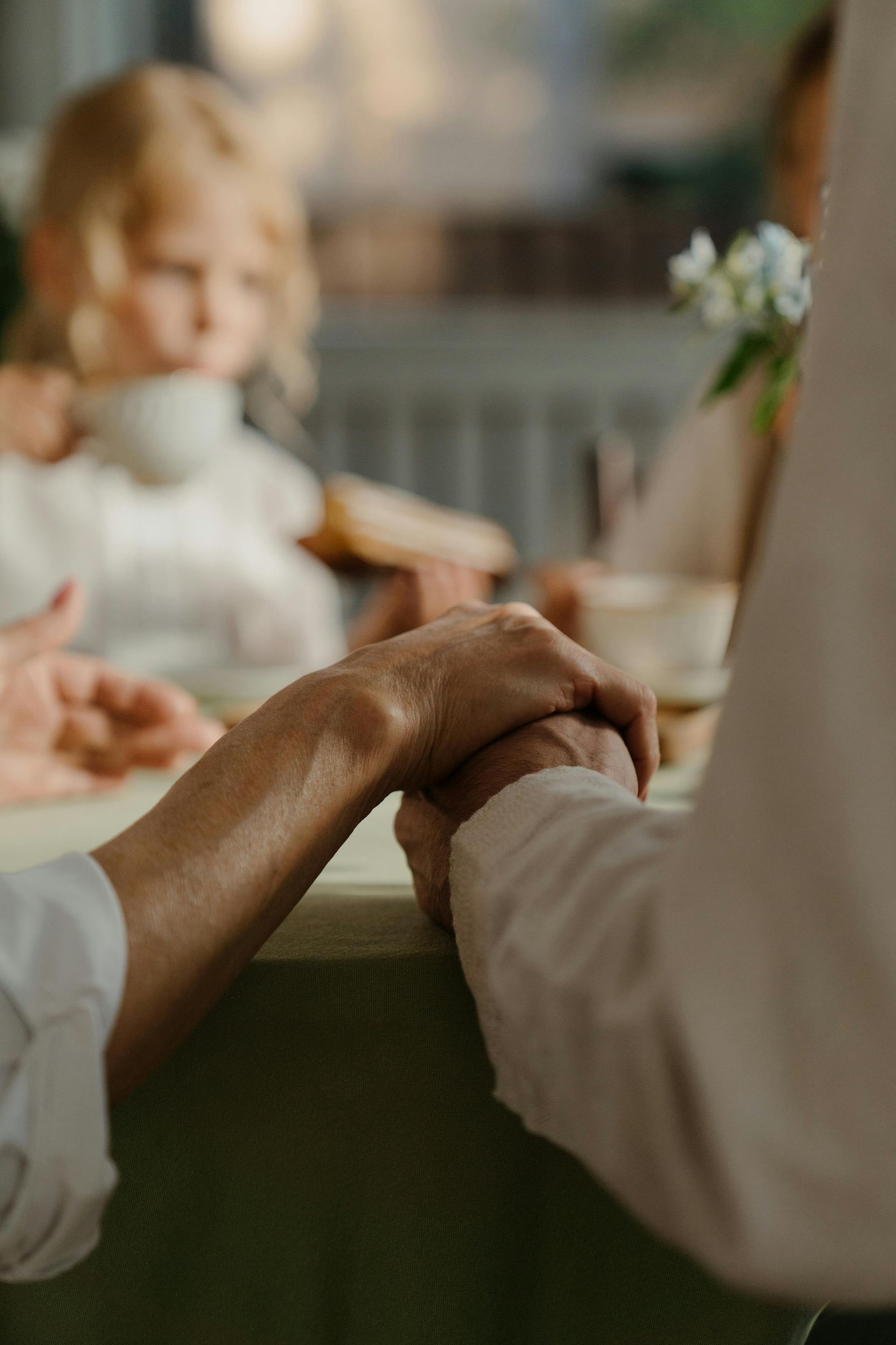 Two people holding hands over a table; a child and another person are blurred in the background.
