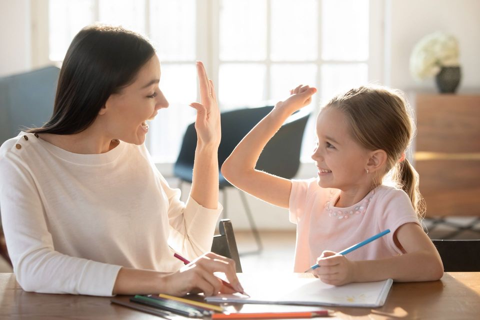 Lehrerin und Kind geben sich an einem Schreibtisch ein High-Five und lächeln in einem hellen Raum mit Papier und Buntstiften.