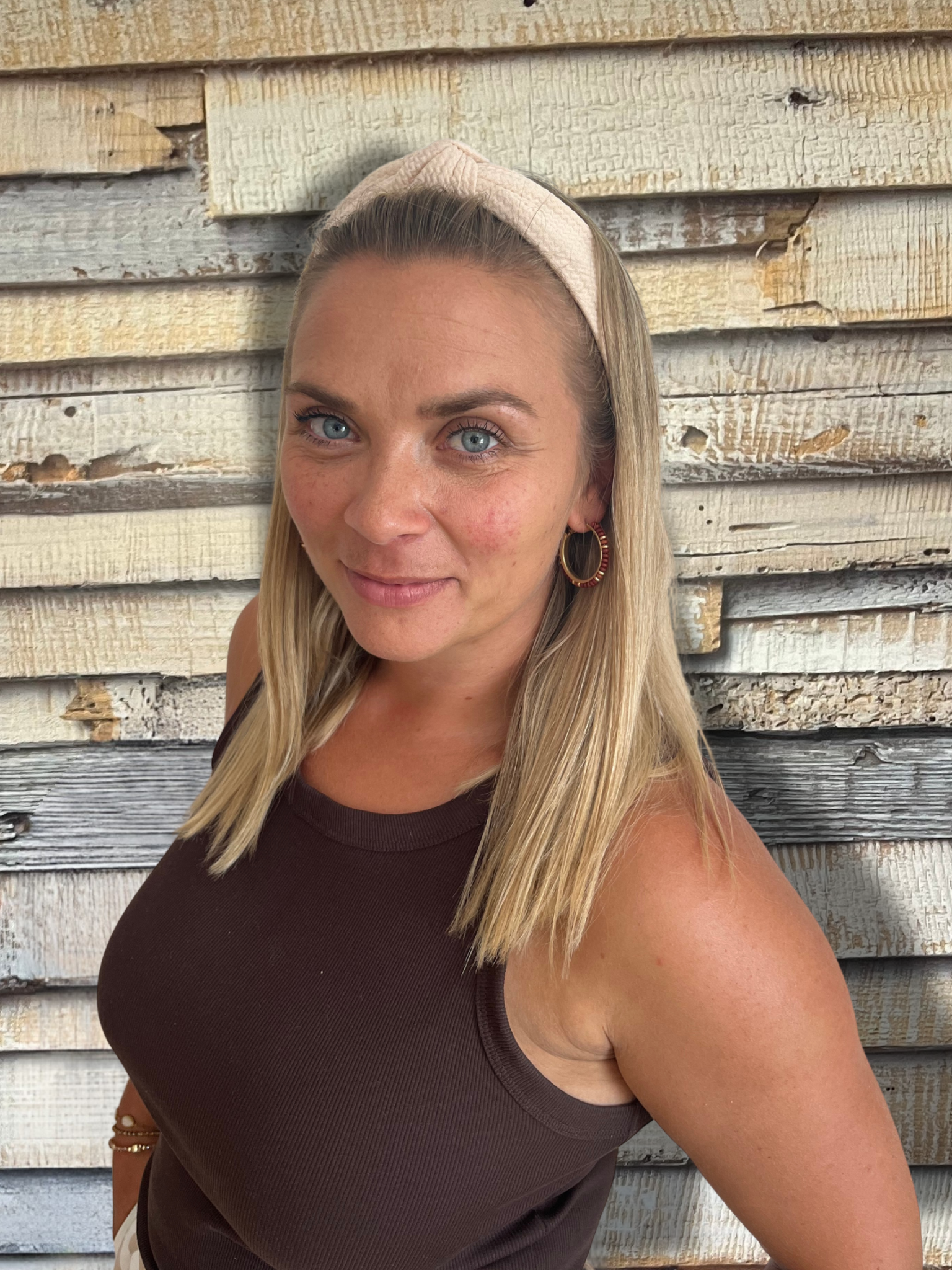Blonde Frau in schwarzem Oberteil und mehreren Halsketten steht vor einer strukturierten Holzwand und lächelt leicht.