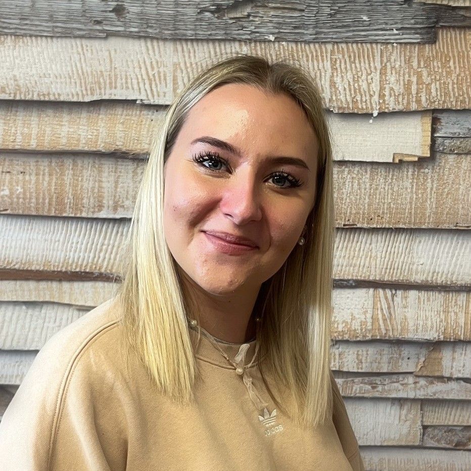 Eine lächelnde blonde Frau in einem beigen Pullover posiert vor einer strukturierten Holzwand.