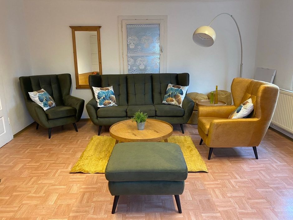 Gemütliches Wohnzimmer mit grünem Sofa, senffarbenem Stuhl, Sessel, Hocker und gelbem Teppich.