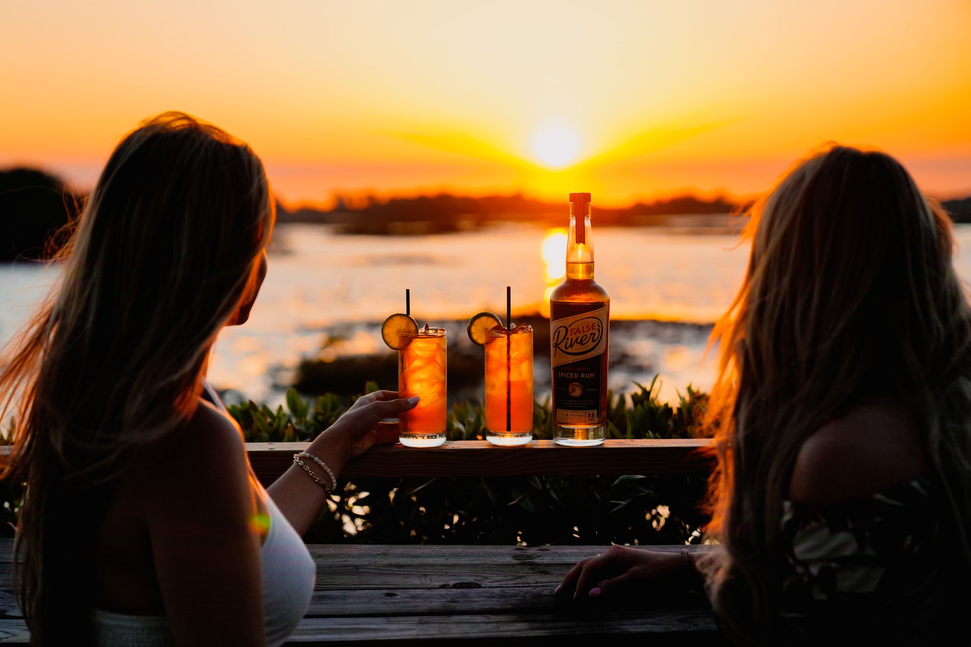 Two women are sitting at a table with drinks and a bottle of alcohol.