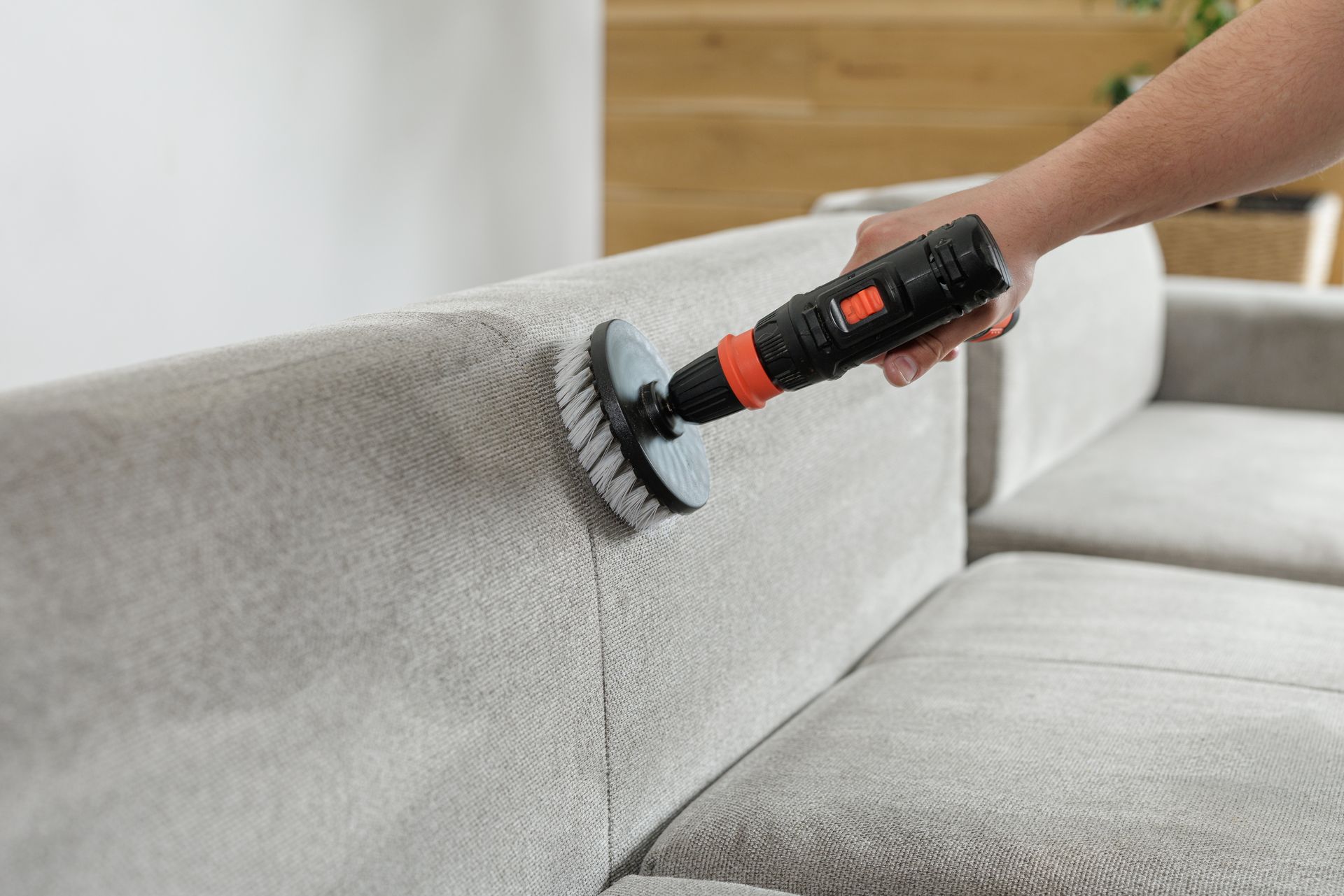 A hand uses a brush vacuum to clean a gray sofa seam.