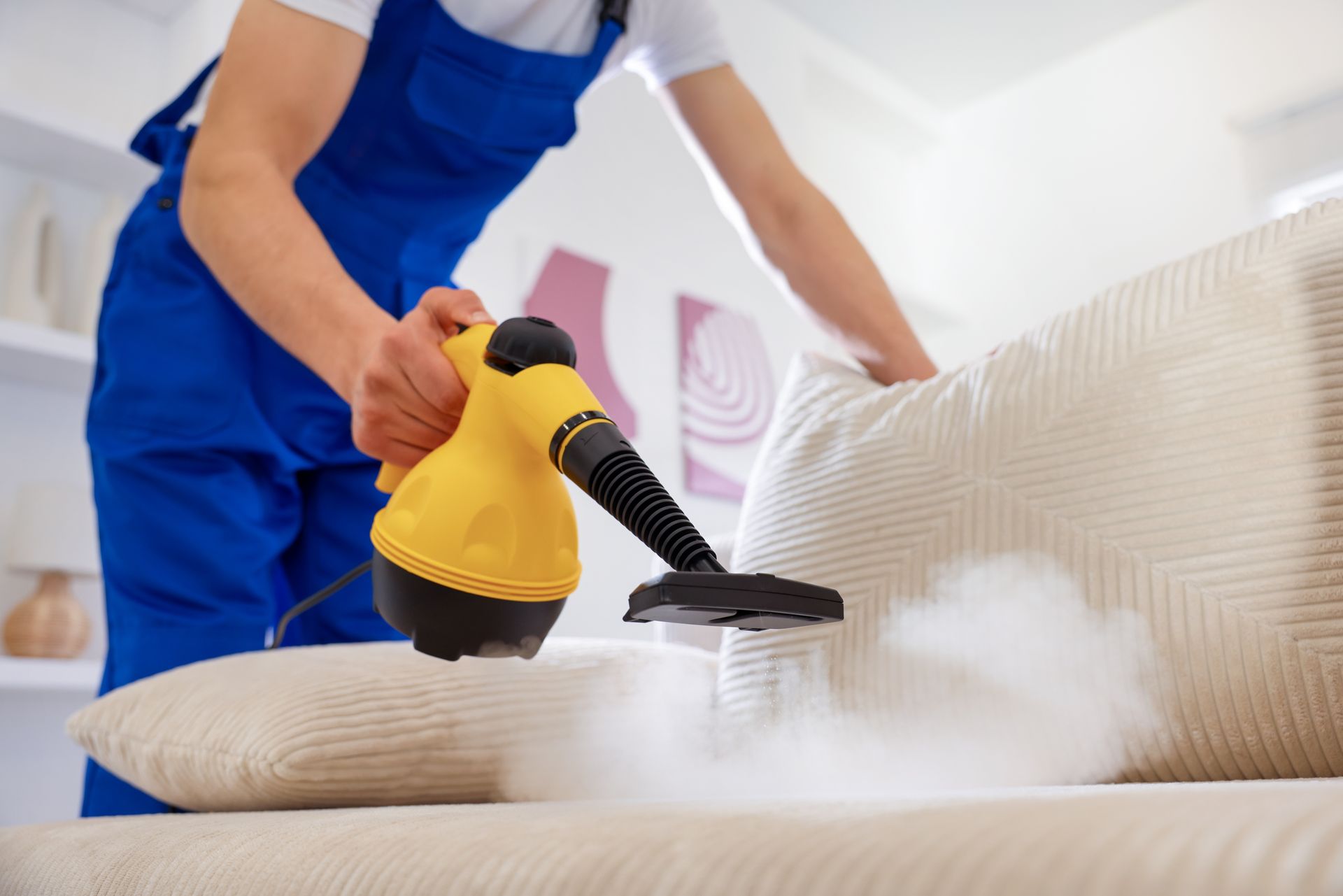Person steam-cleans a beige couch with a yellow handheld cleaner.