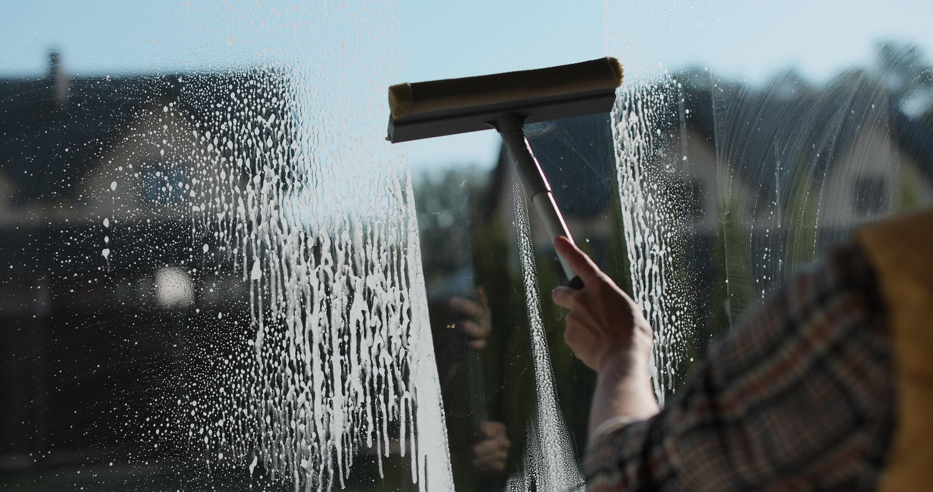 Person using a squeegee to clean a window with water droplets in bright daylight