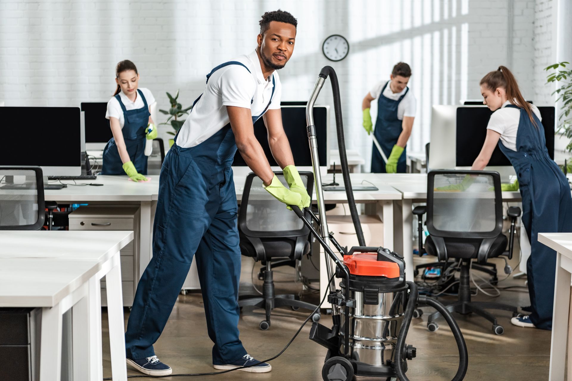 People cleaning a bright office, with one vacuuming in the foreground and others tidying desks.