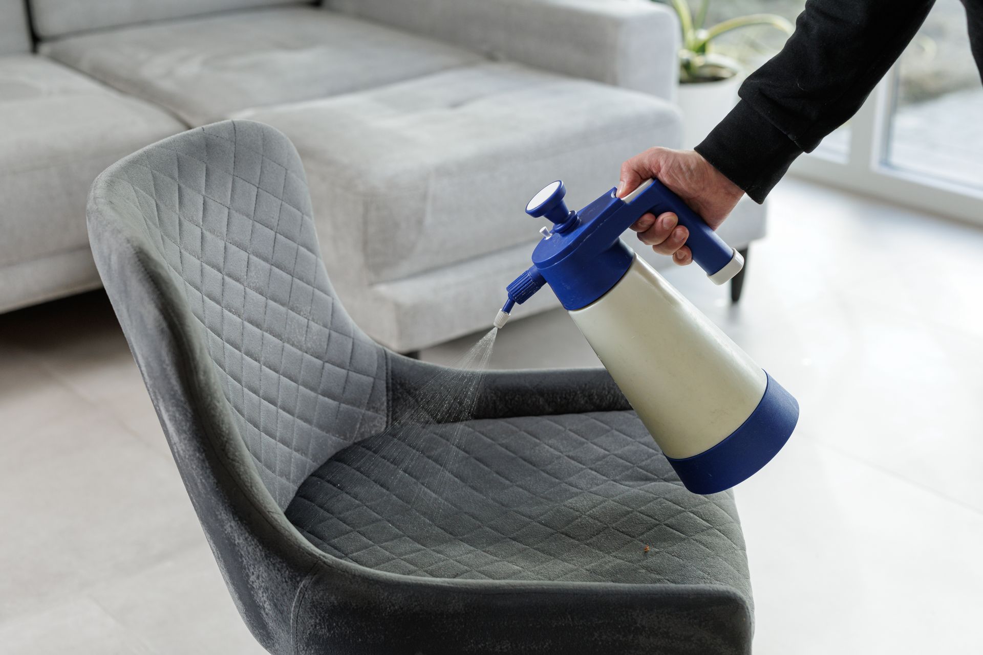 Hand spraying cleaner on a gray upholstered chair in a bright living room