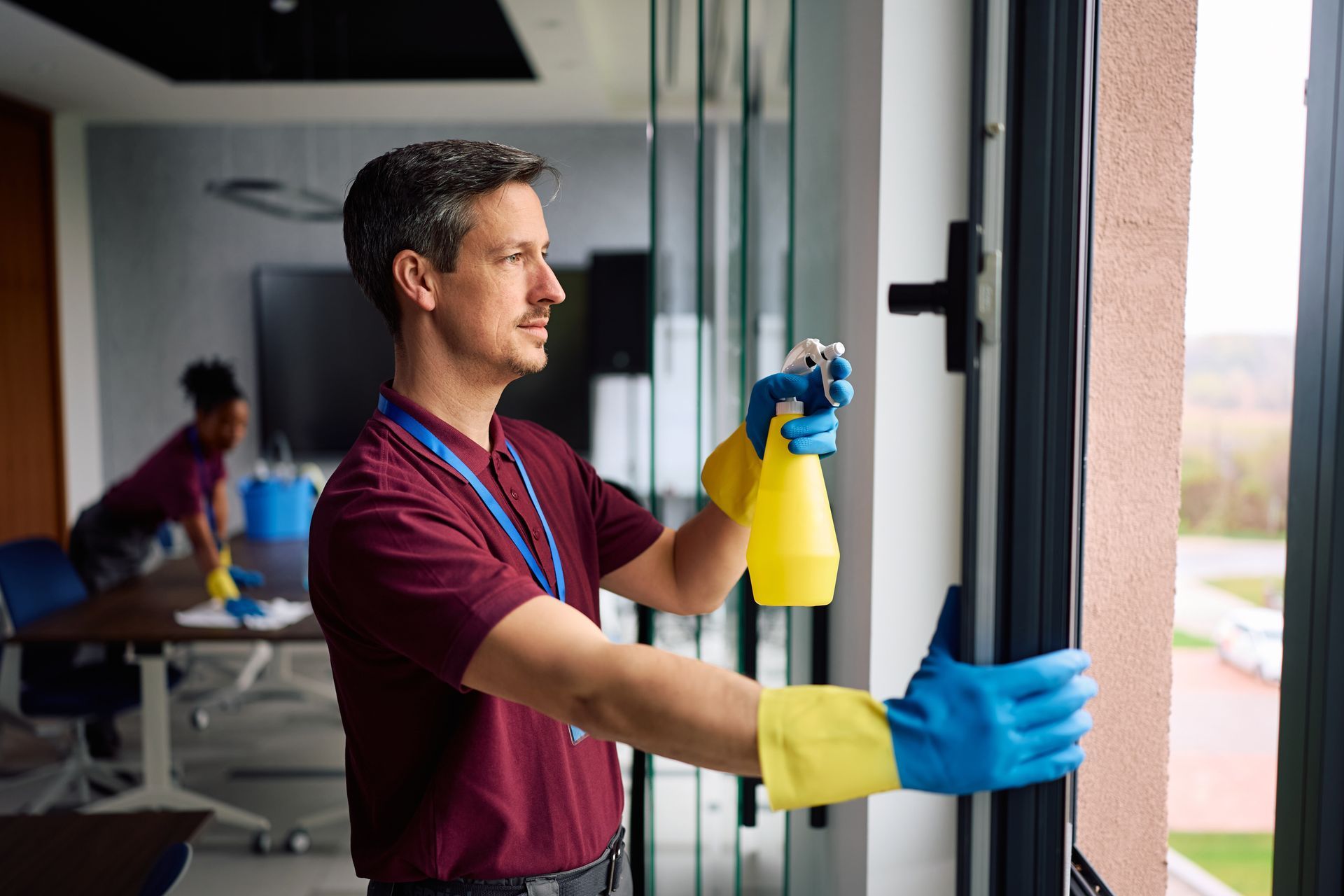 Worker in blue gloves sprays cleaner on a glass window inside an office.
