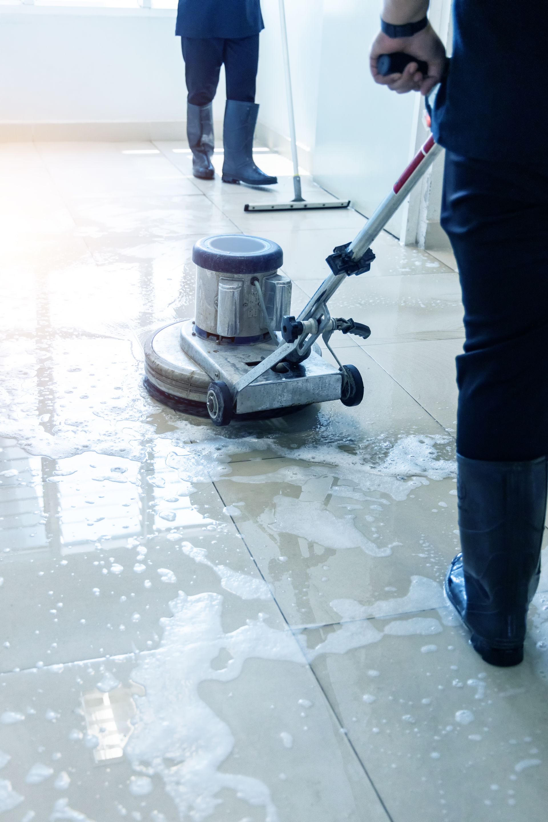 Floor buffer cleaning a wet, soapy tile floor with two workers nearby