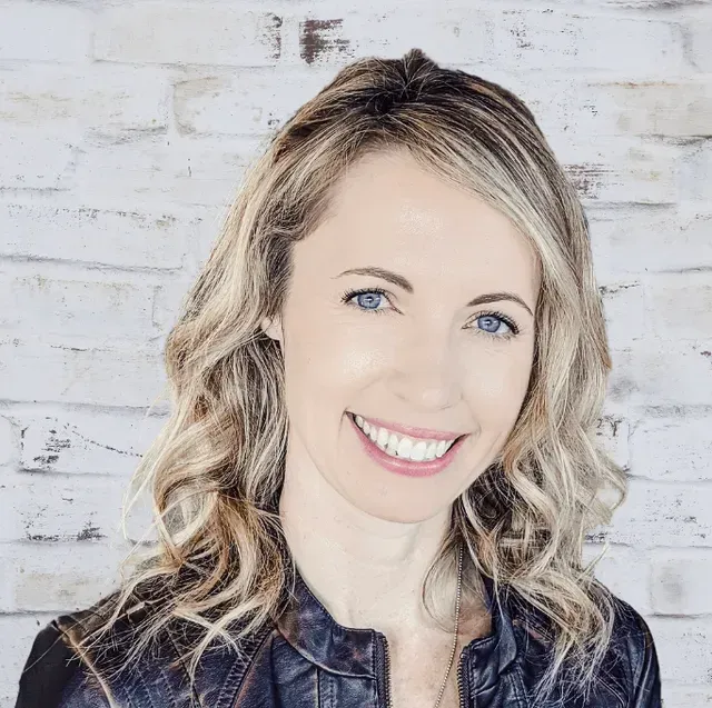 Woman with blonde hair smiles at the camera against a white brick wall. She wears a leather jacket.