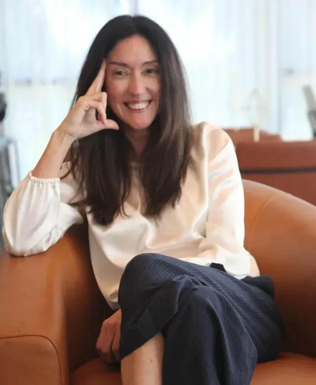 Woman in white blouse and blue pants smiles while sitting in a brown chair.