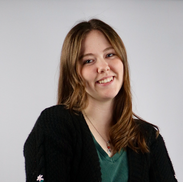 Woman with long brown hair, smiling, wearing a black sweater over a green shirt, against a gray background.