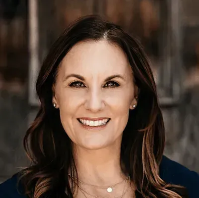 Woman with brown hair smiles, wearing a navy top, outside a wooden structure.