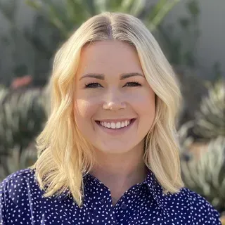 Blond woman smiling, wearing a blue polka-dot shirt, with a background of greenery.