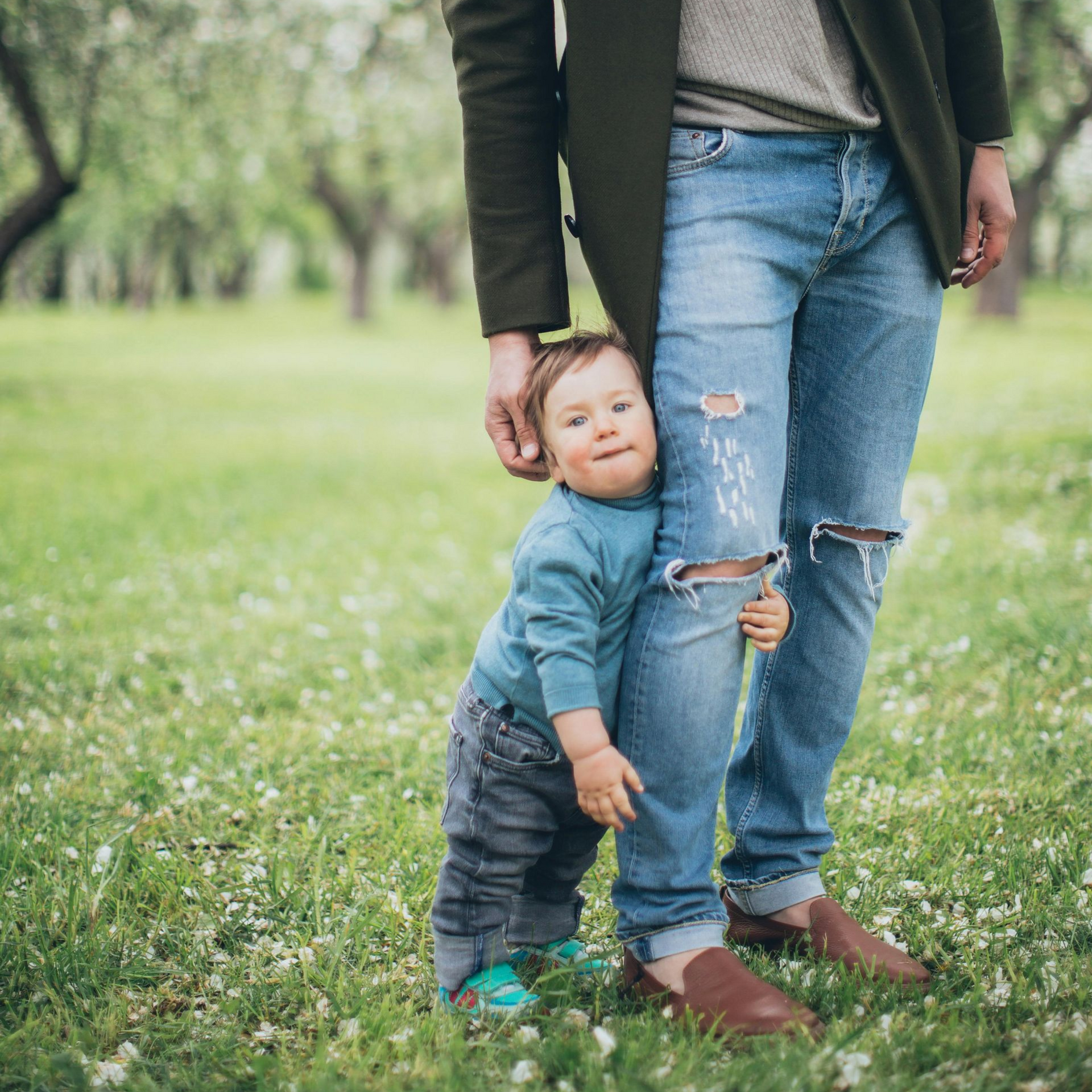Toddler hugging an adult’s leg in a grassy park, wearing a blue sweater and gray pants at Ava's Path