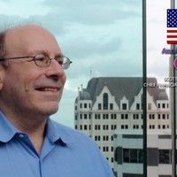 Man wearing glasses smiles, with urban cityscape in background, American flag in frame.