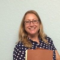 Woman with glasses smiling, holding brown folder, wearing a navy polka dot shirt, against a pale blue wall.