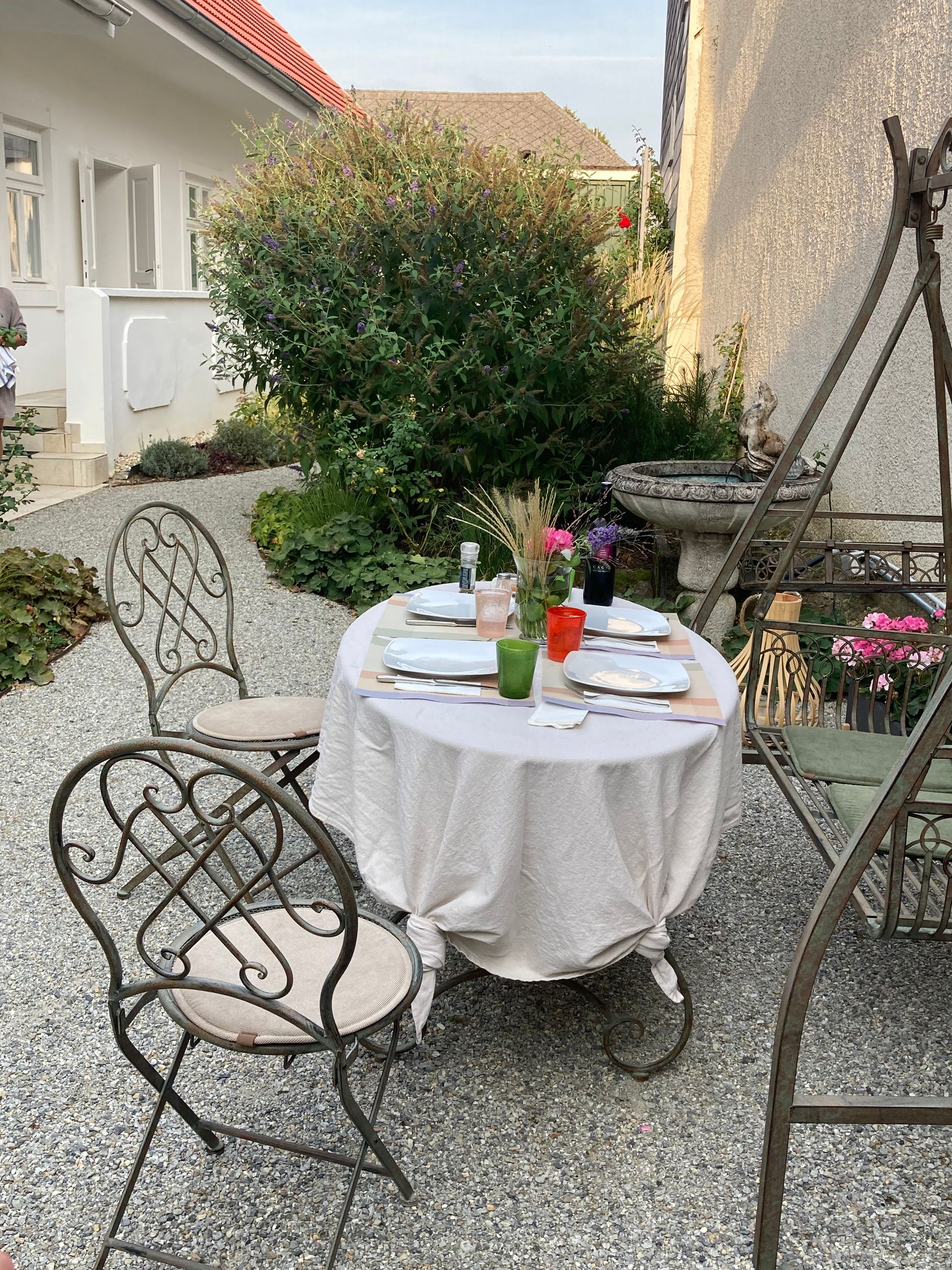 Essgruppe im Freien: gedeckter Tisch, verzierte Stühle und eine Schaukel in einem Garten mit Kiesboden.