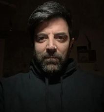 Un uomo con la barba indossa una felpa con cappuccio nera e guarda la telecamera.