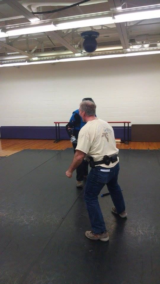 A man in jeans and a tan shirt practices martial arts with another person in a gym.