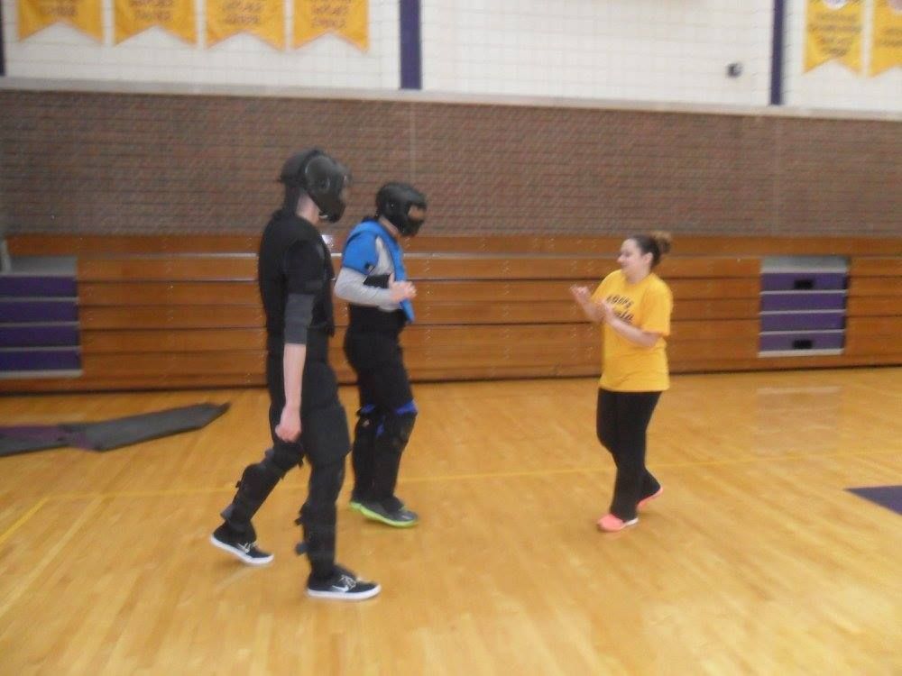 Three people in a gym; two in combat gear, facing a woman in yellow. The woman gestures with her hands, likely instructing.