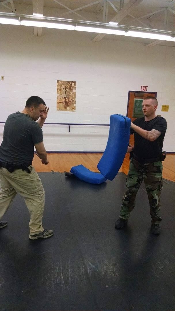 Two men in tactical gear practice self-defense; one punches a blue padded object held by the other in a gym.