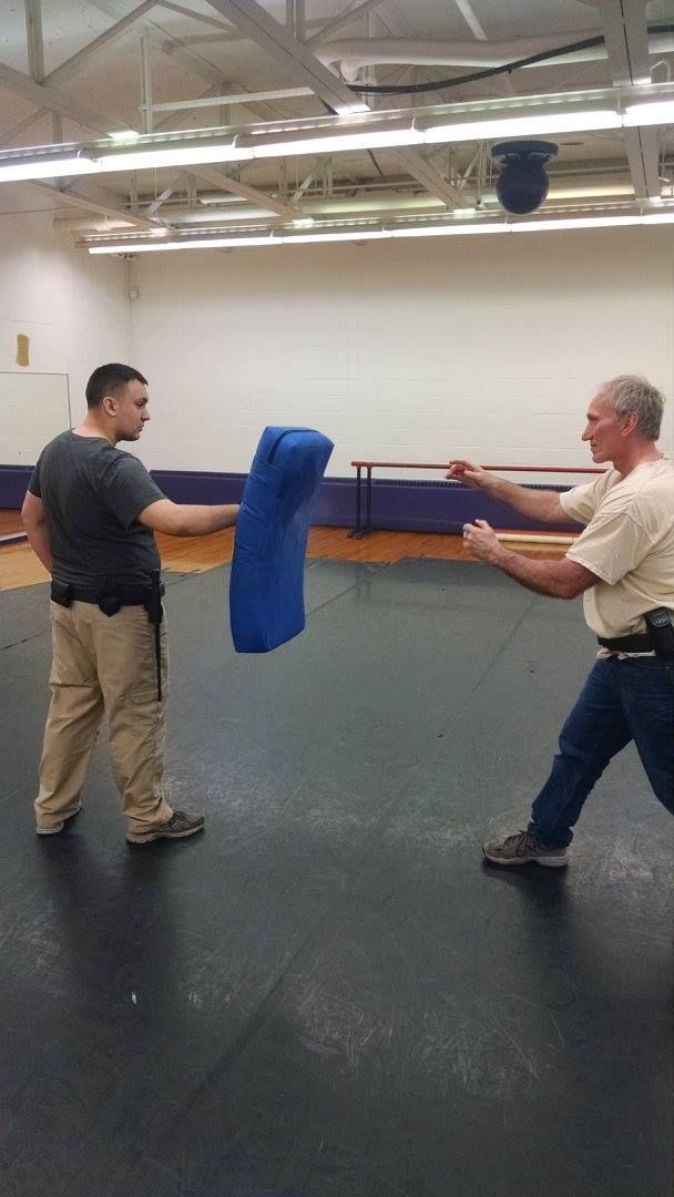 Two men practicing self-defense in a gym. One holds a blue padded target, the other throws a punch.
