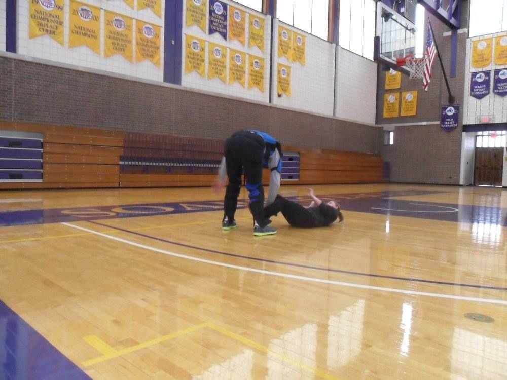 Person helping another person on the ground in a gymnasium. One person is bent over, the other is lying down.