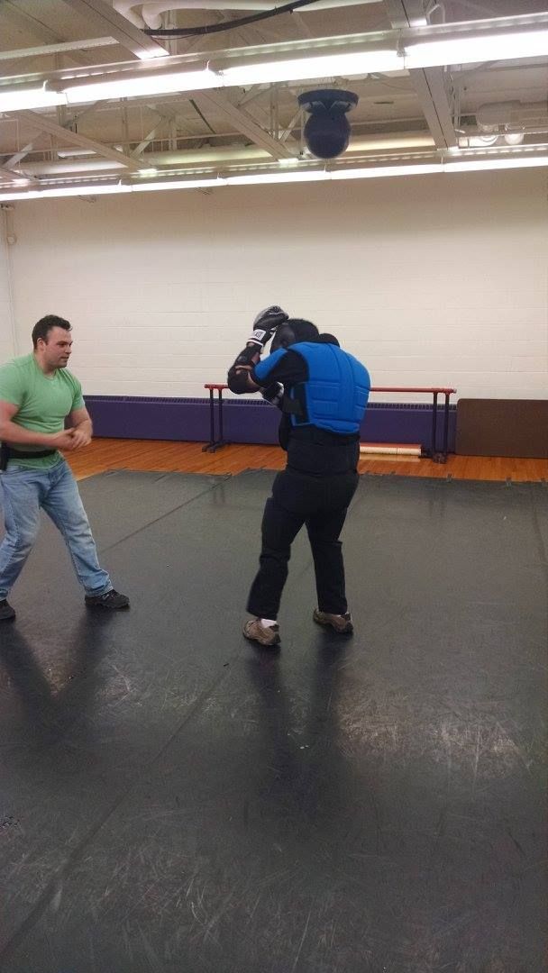 A man in a green shirt and jeans faces a person wearing protective gear in a gymnasium. The person is crouched.