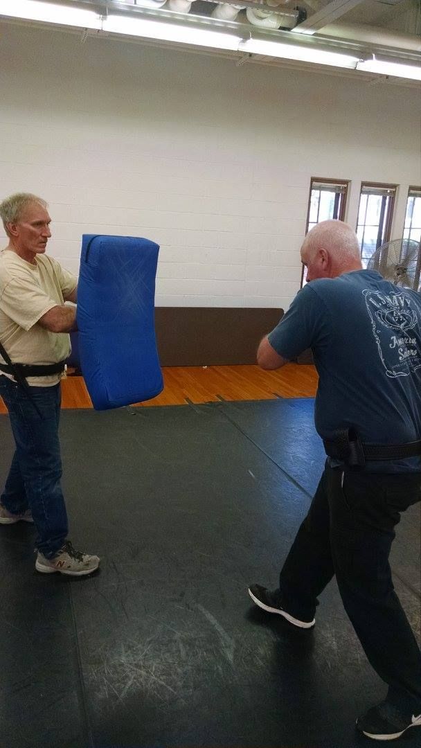 Two men in a training room: one holding a blue pad, the other kicking at it.