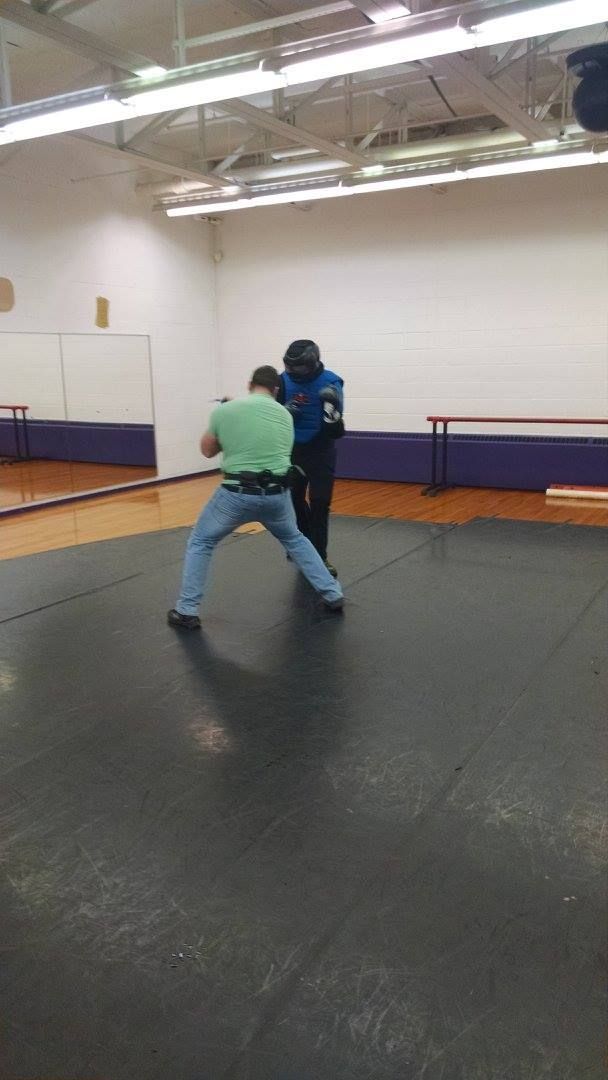 A man in jeans and a green shirt practices with a person wearing a blue padded suit in a gym.